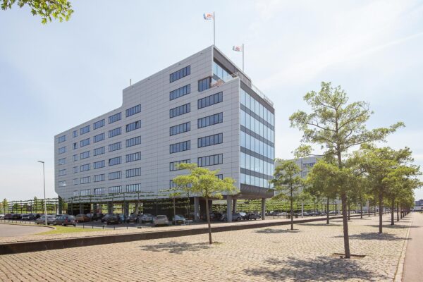 Modern kantoorgebouw aan de Waalhaven Oostzijde in Rotterdam met parkeerruimte eronder en een met bomen omzoomde straat.